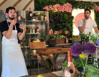 Two men presenting at an elegant outdoor event with flowers and champagne